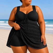 A black one-piece swim dress with a structured top, wide straps, and a flowing skirt overlay featuring a textured pattern.